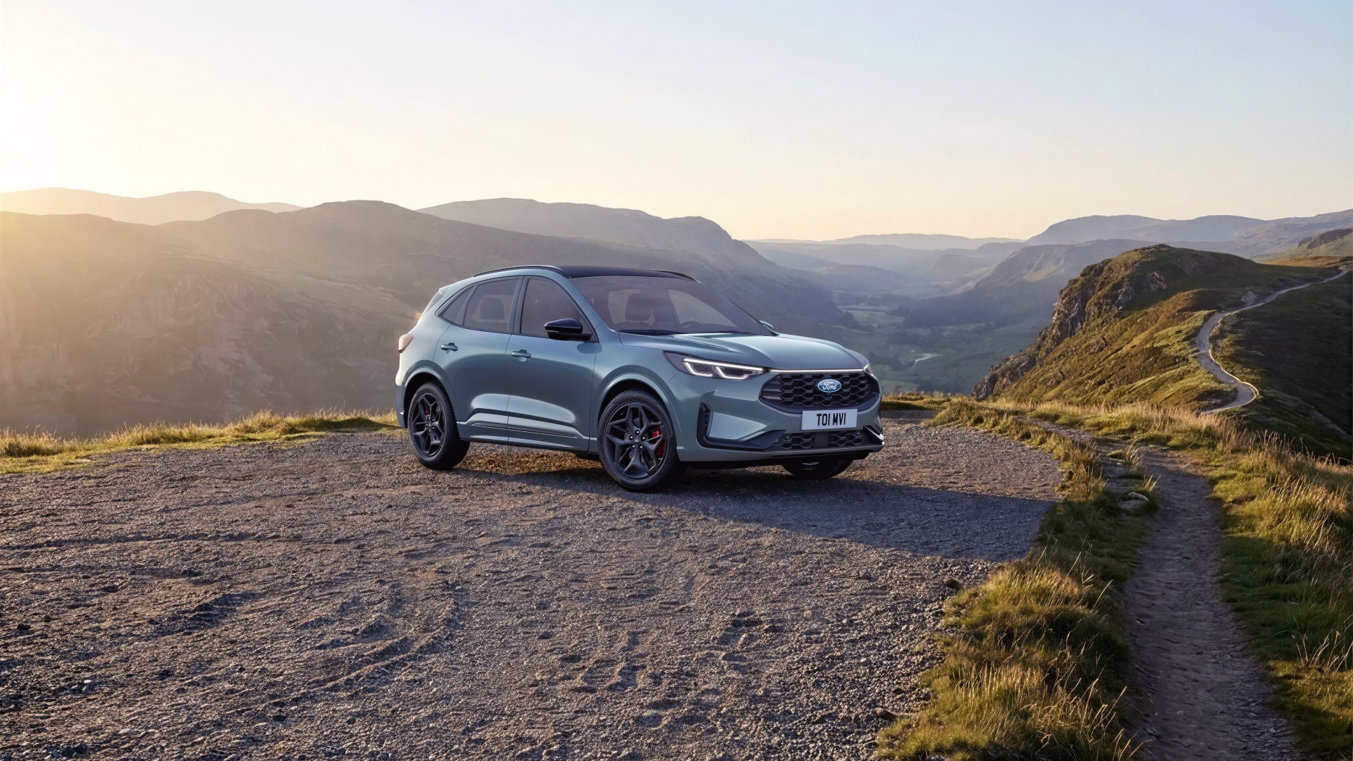 Un Ford Kuga grigio è parcheggiato su un sentiero sterrato che si affaccia su un paesaggio montuoso con una strada tortuosa.