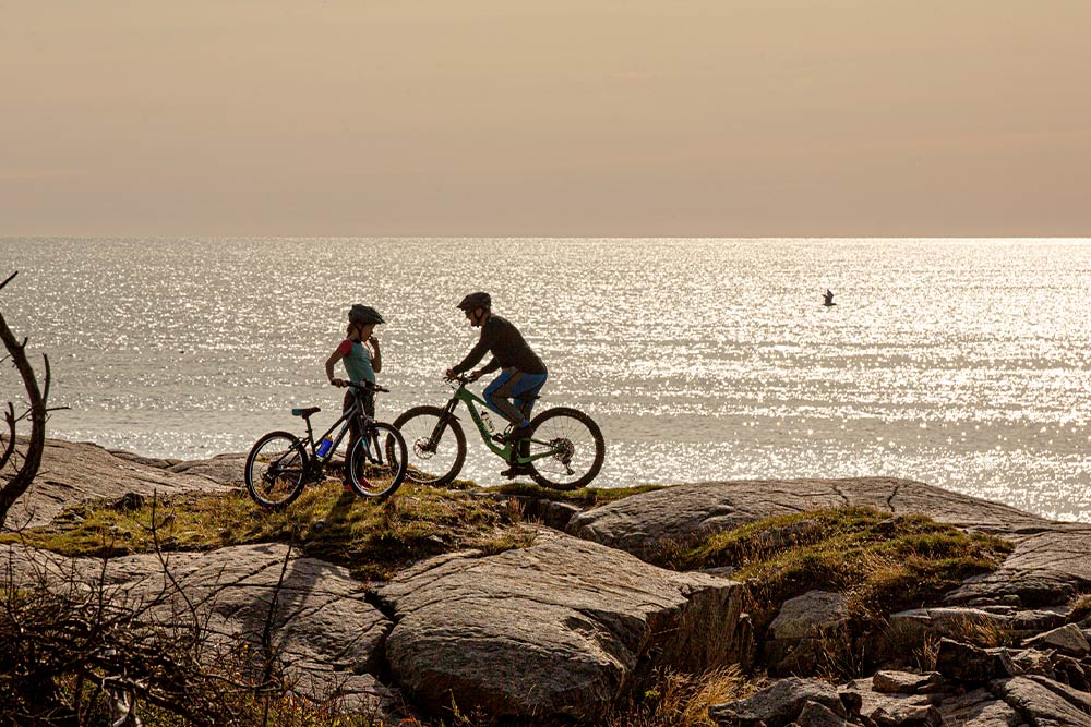 Due persone in mountain bike su una riva rocciosa, che guardano il mare illuminato dal sole. 
