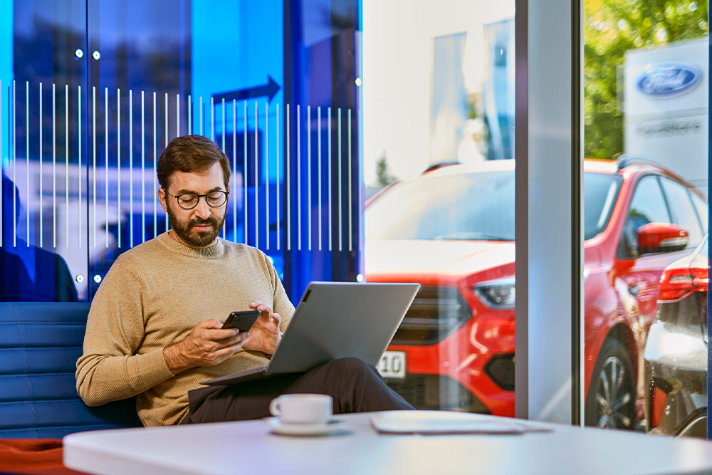 Persona seduta in una sala d'attesa di una concessionaria Ford, guarda il telefono con un laptop vicino.