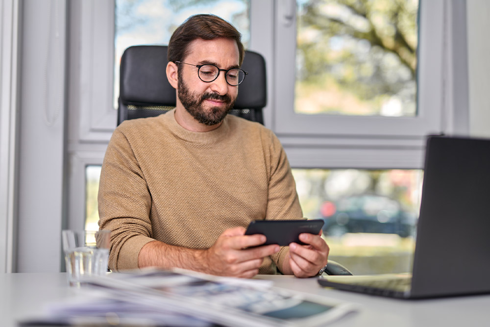 Persona seduta a una scrivania, guarda uno smartphone che tiene in mano, con un laptop sulla scrivania.