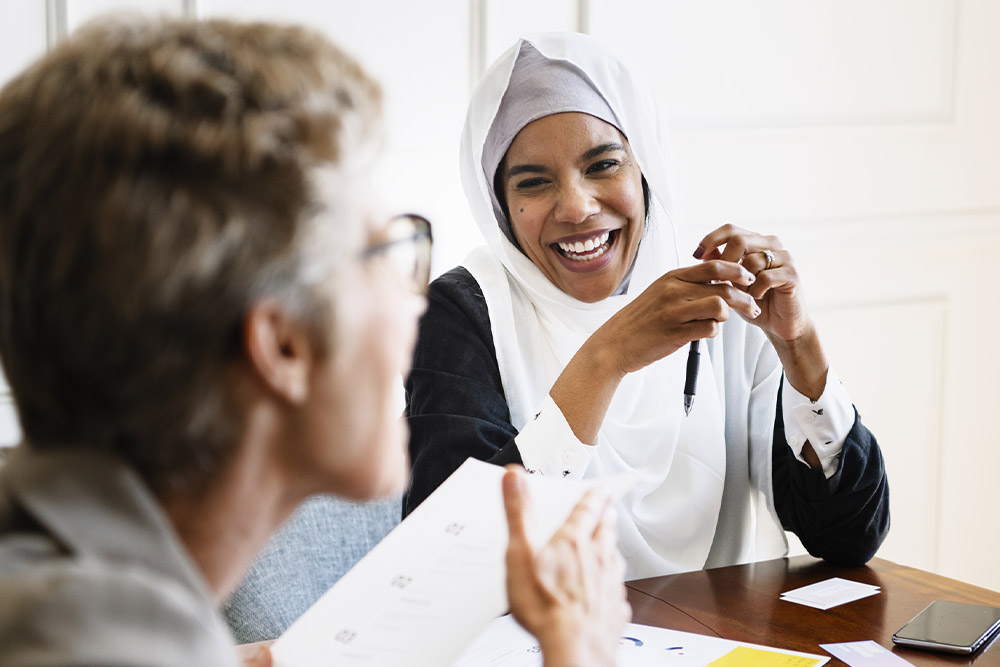 Una donna con un hijab bianco sorride durante una riunione, tenendo una penna. Documenti e un telefono sono sul tavolo.