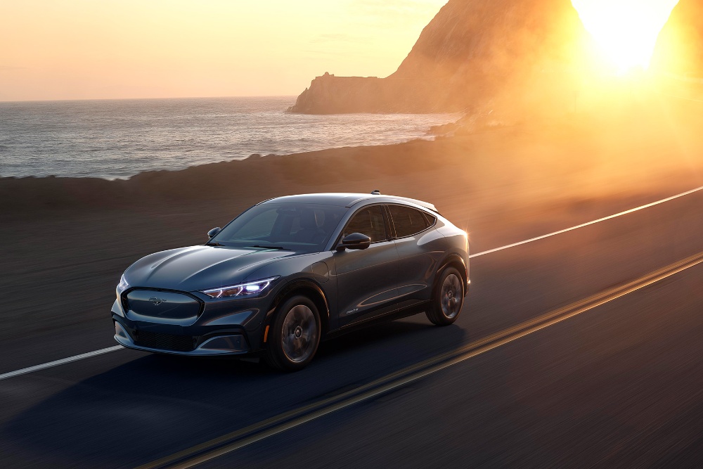 Un'auto elettrica Ford Mustang Mach-E percorre una strada costiera al tramonto.