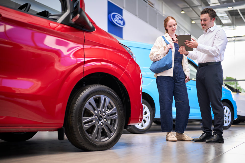 Due persone in uno showroom Ford guardano un tablet, con un furgone Ford rosso in primo piano e un furgone Ford blu dietro.