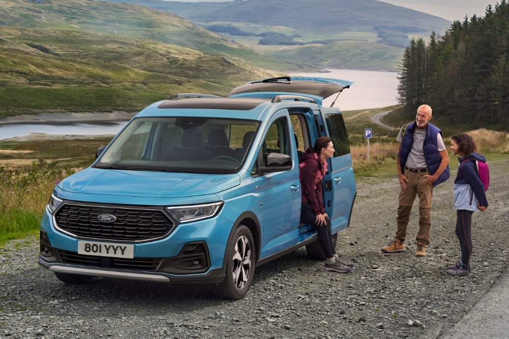 A blue Ford Tourneo Connect parked by a lake, with people getting out of the vehicle.