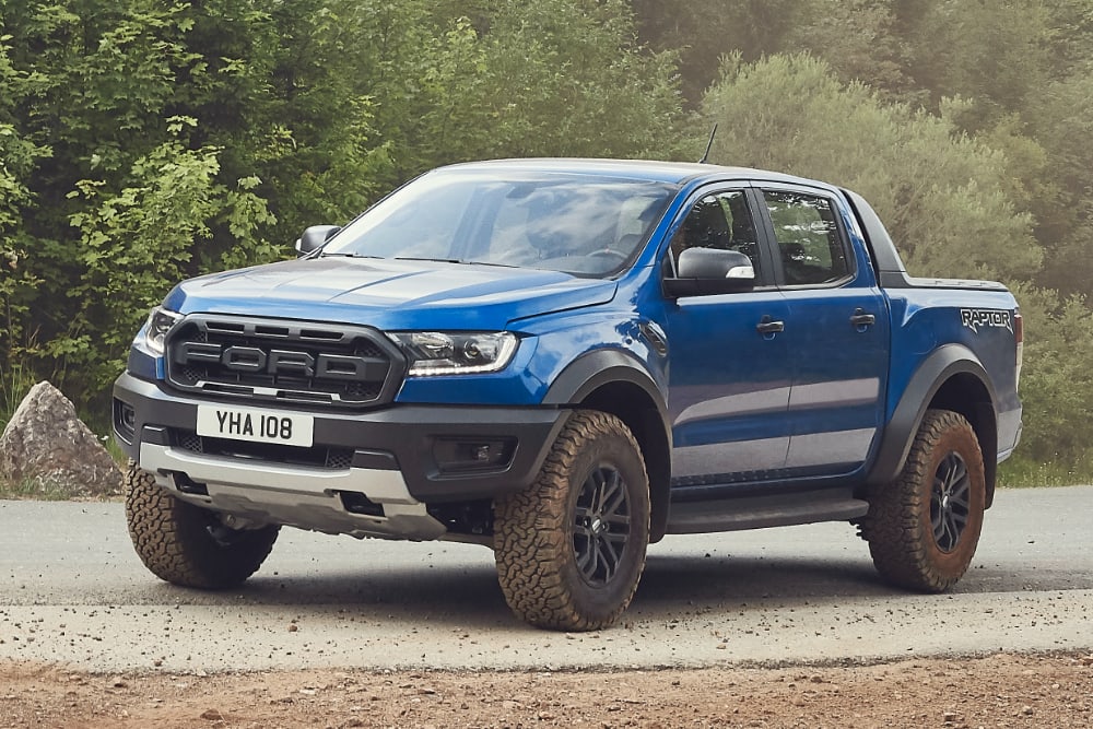 A blue Ford Ranger Raptor pickup truck on a dirt road, surrounded by trees.