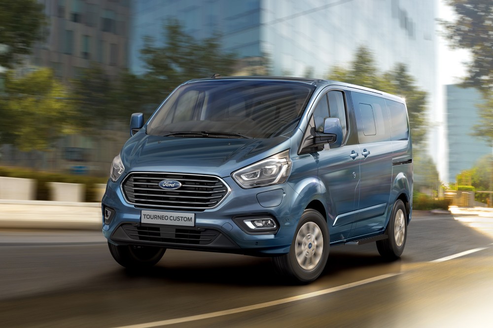 Front 3/4 view of a blue Ford Tourneo Custom driving on a city street.
