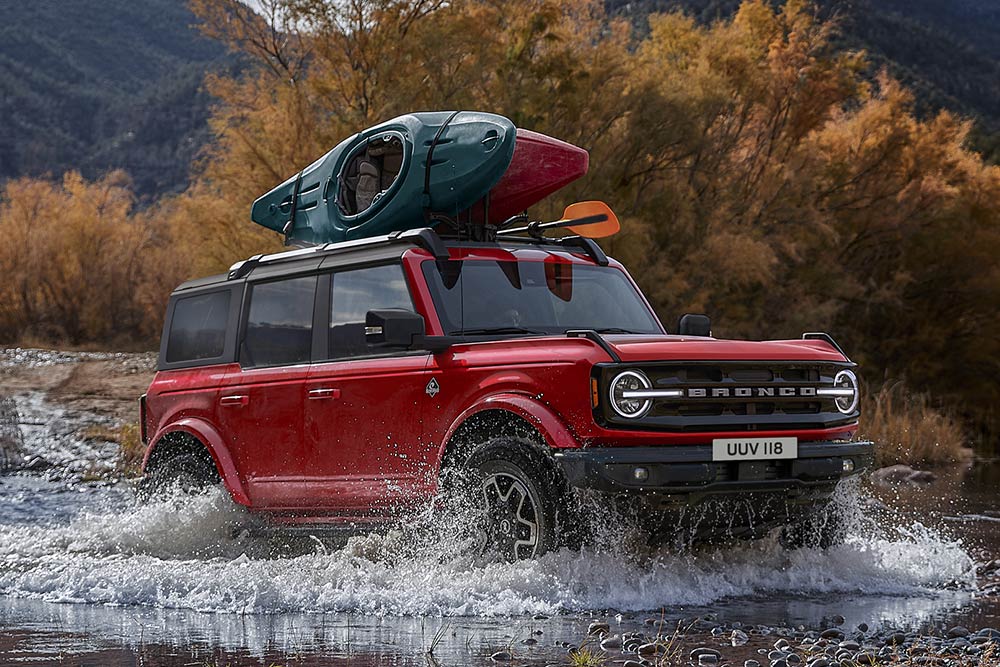 Vista laterale di un Ford Bronco rosso che attraversa un corso d'acqua non profondo in montagna.