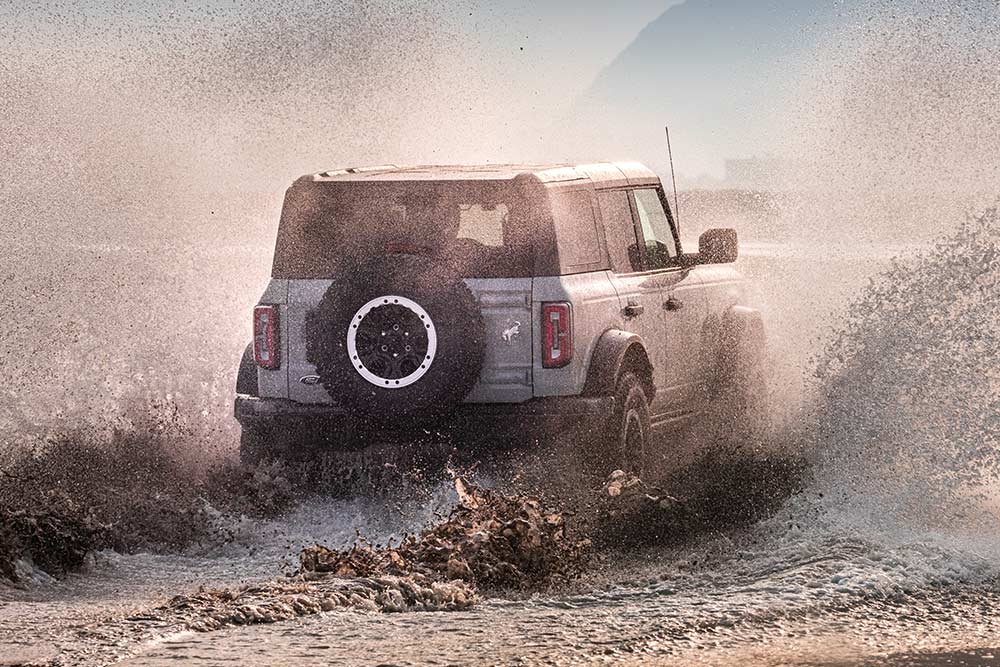 Vista posteriore di un Ford Bronco color argento che attraversa una spiaggia alzando uno schizzo d'acqua.