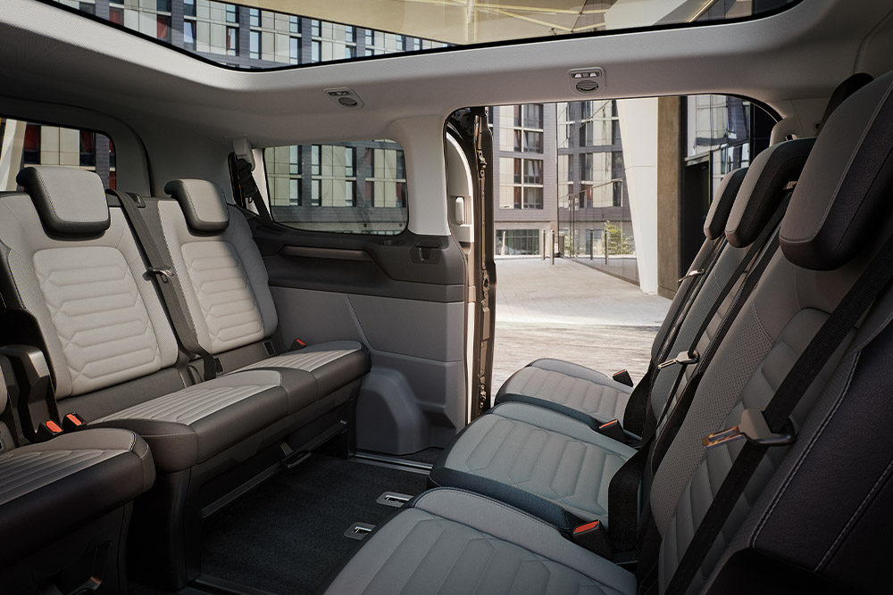 Vista sugli interni del Ford Tourneo Custom che mostra le file dei sedili dei passeggeri grigi e il tettuccio panoramico.