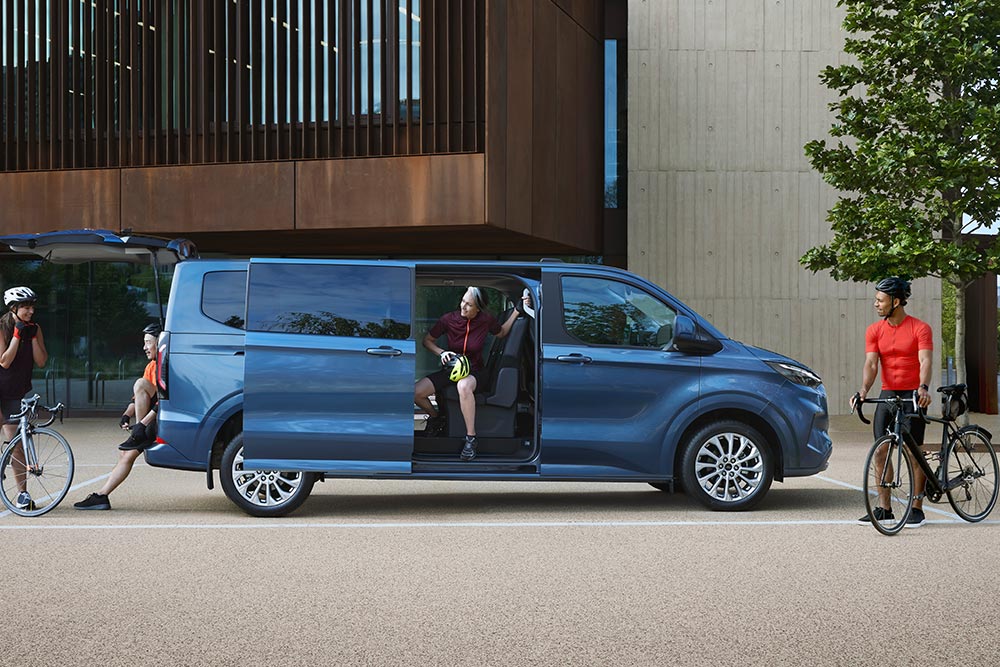 Un furgone Ford Tourneo Custom blu parcheggiato con le portiere laterali aperte e alcune persone in bicicletta nelle vicinanze. 