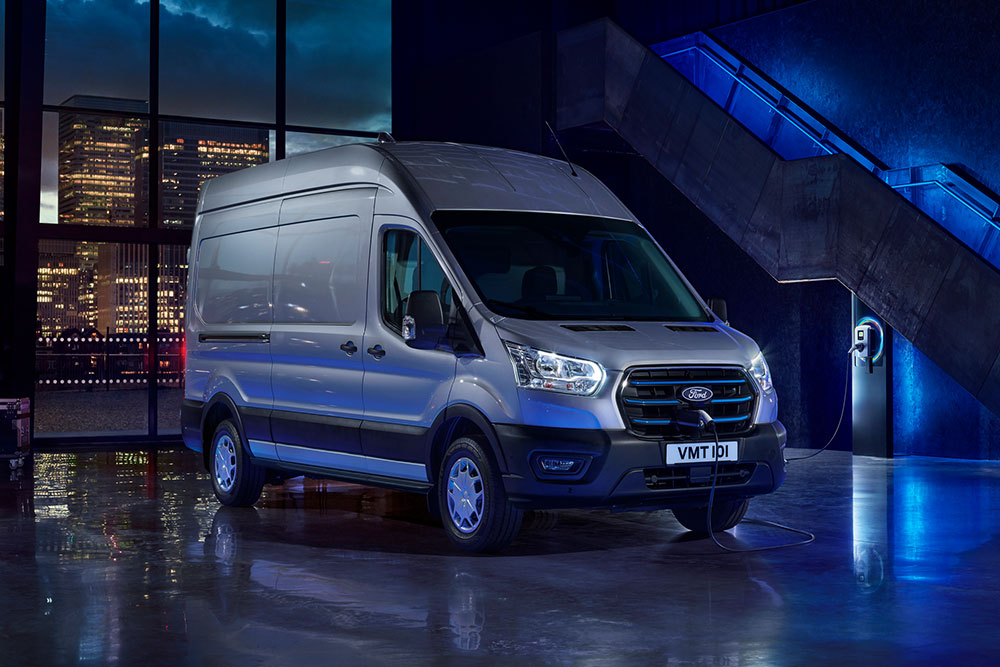 Un furgone elettrico E-Transit in versione argento parcheggiato al chiuso con una città visibile sullo sfondo attraverso grandi vetrate. 
