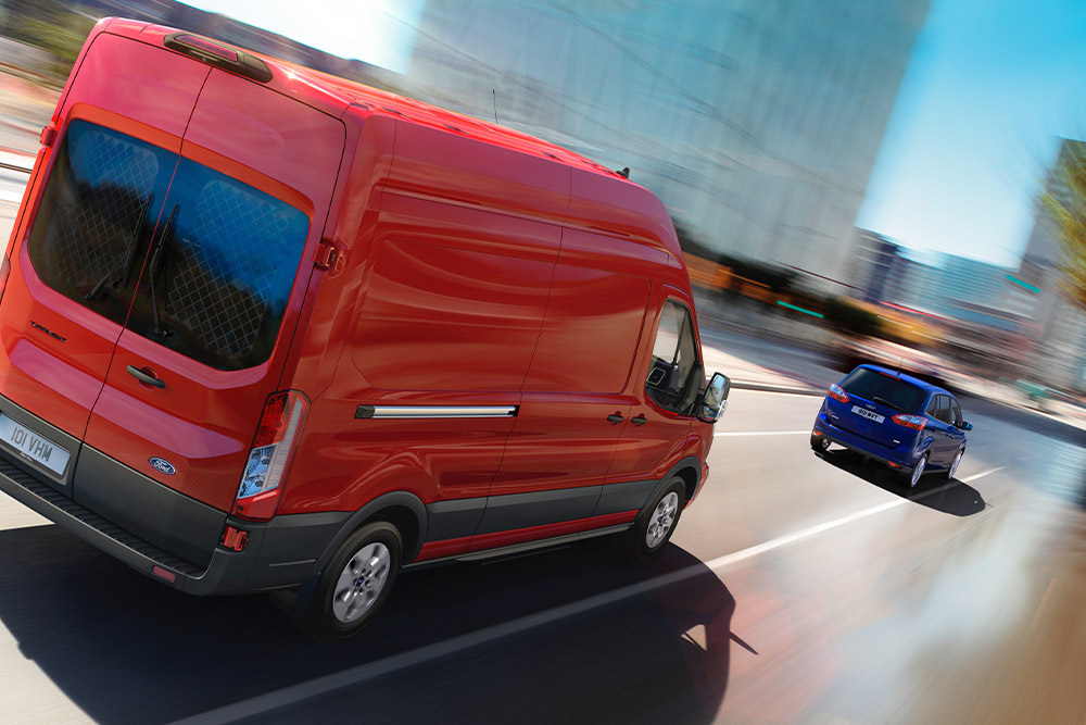 Rear 3/4 view of a red Ford Transit driving on a city road, with a blue car ahead.