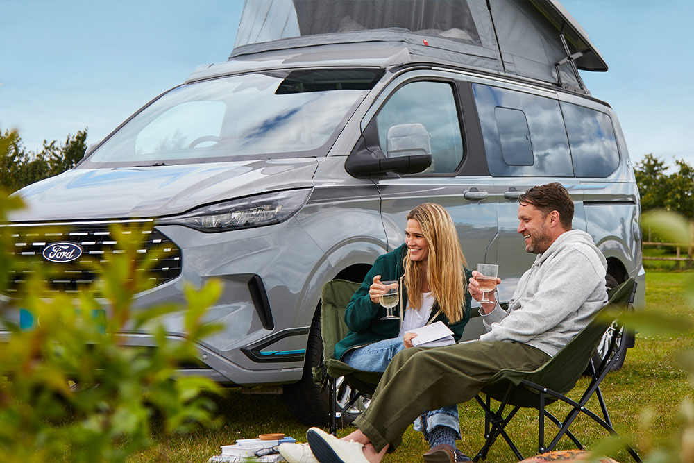 Una coppia che si gode un drink su sedie da campeggio accanto a un camper Ford Transit Custom grigio con tetto a soffietto.