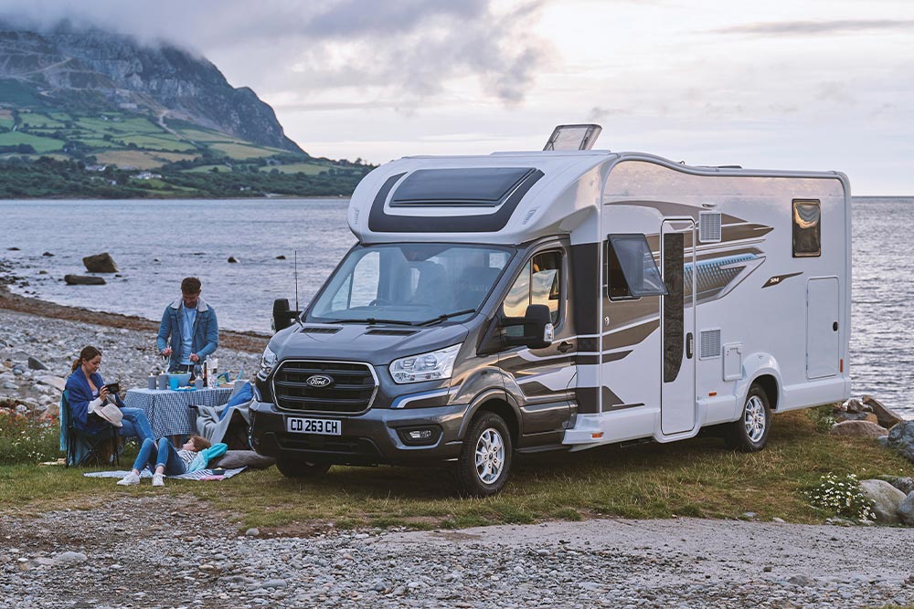 Una coppia che si gode un drink su sedie da campeggio accanto a un camper Ford Transit Custom grigio con tetto a soffietto.