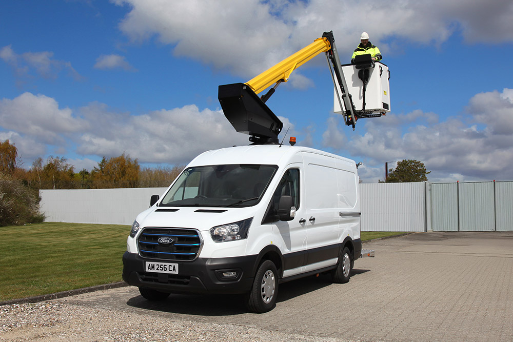 Un furgone Ford Transit bianco con una piattaforma aerea estesa, un tecnico nel cestello, sotto un cielo blu.