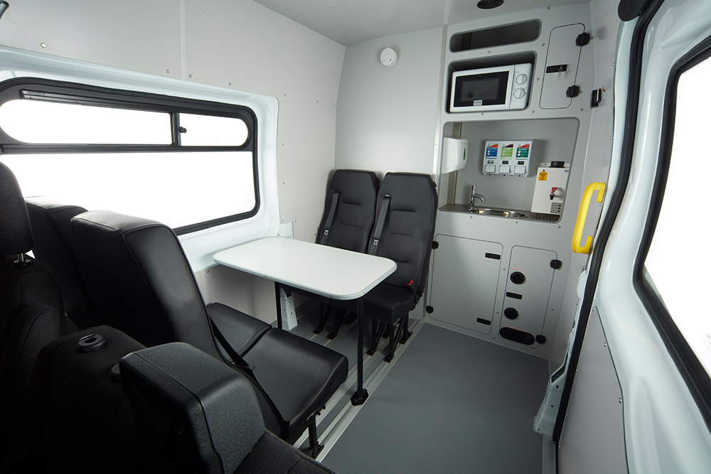 Interno di un furgone Ford, con tavolo, sedili neri e un angolo cottura compatto con microonde.