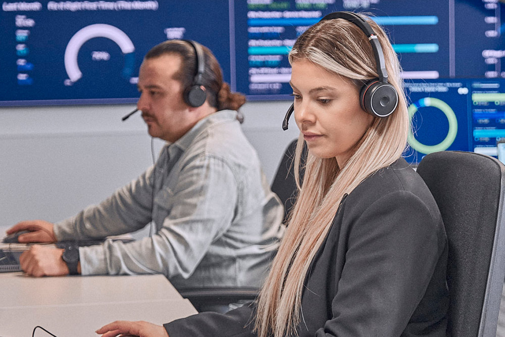 Due persone con cuffie concentrate su schermi di computer in una sala di controllo moderna, con display dati.