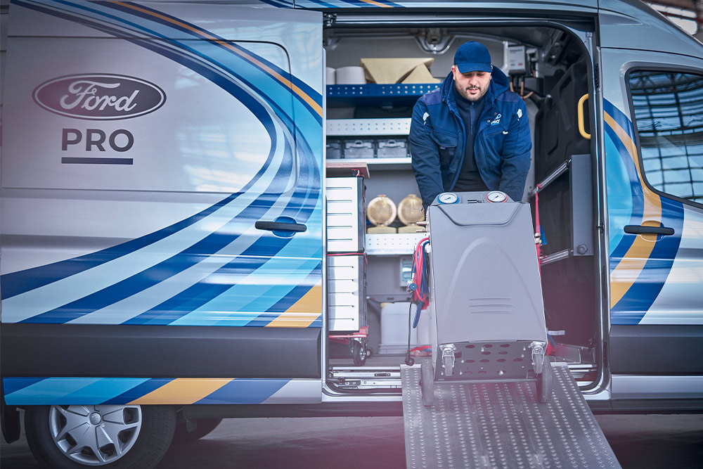 Un tecnico mobile Ford Pro est intento a estrarre attrezzi da un furgone Transit davanti a un'azienda.
